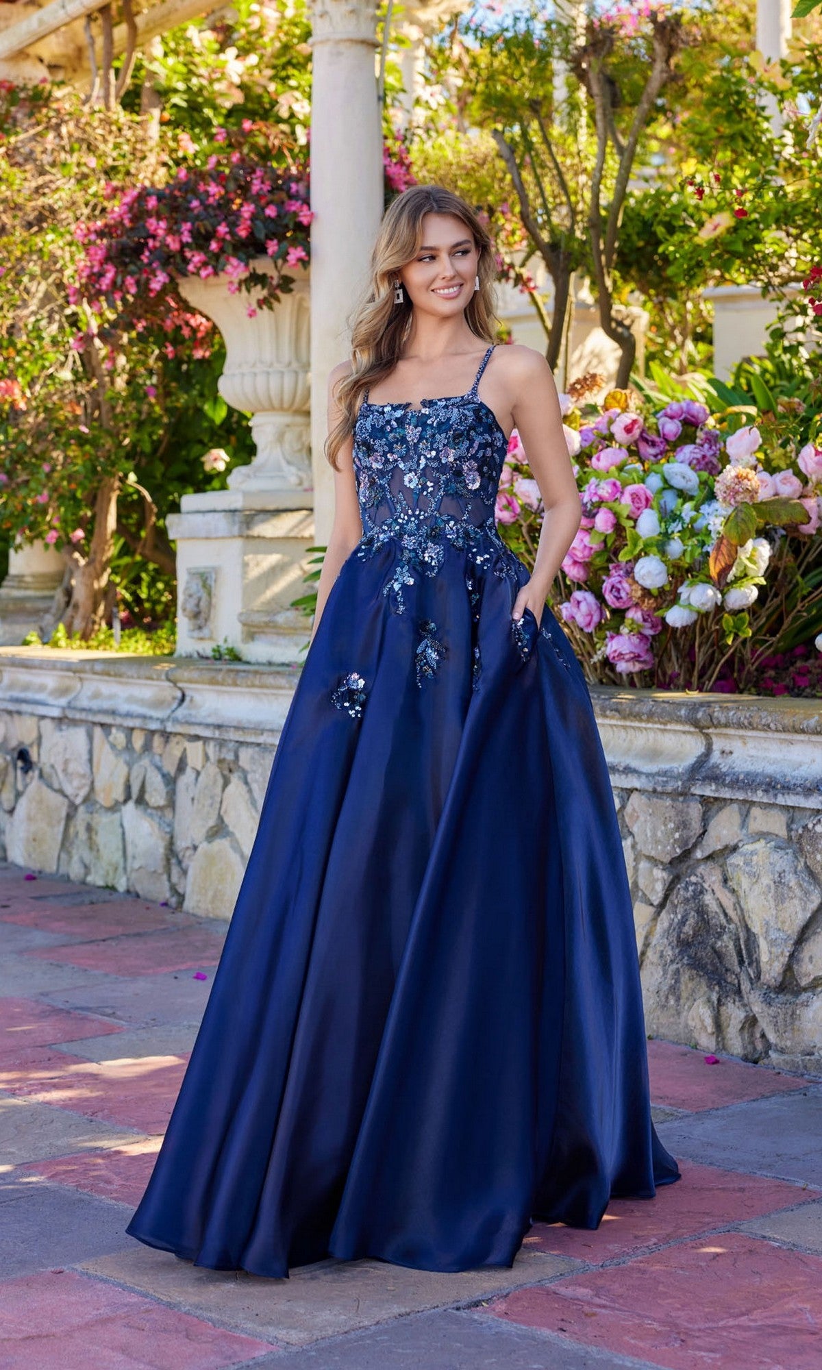 A woman in the Juliet JT2617U long prom dress, featuring navy blue floral sequin embellishments and a lace-up back, stands by a stone wall surrounded by blooming flowers.